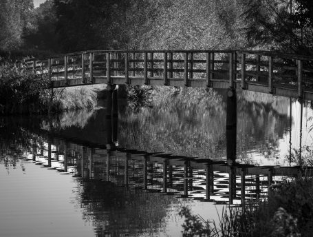 Zwart-wit afbeelding van een houten brug met reflectie in het water.