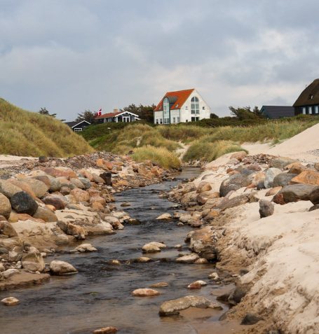 Een beekje stroomt langs een strand met huizen en duinen op de achtergrond.