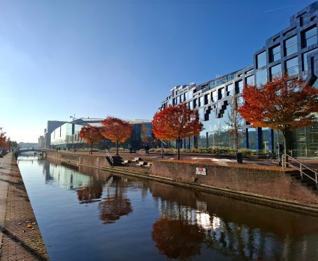 Stadsgracht met herfstbomen en moderne gebouwen onder een blauwe lucht.