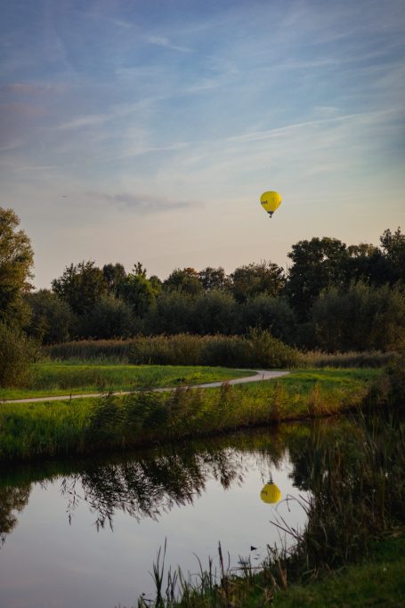 Gele luchtballon boven een groene oever met reflectie in het rustige water.