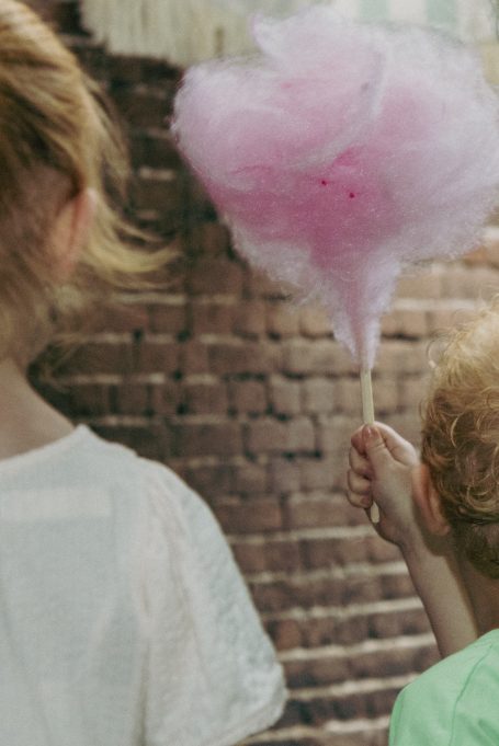 Meisje met een suikerspin staat naast een ander kind, voor een bakstenen muur.