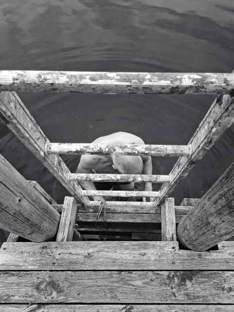 Persoon klimt een ladder naar een wateroppervlak, omringd door houten palen.