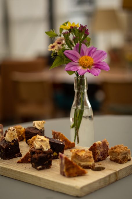 Assortiment van gebakjes op een plank met een vaasje bloemen op tafel.